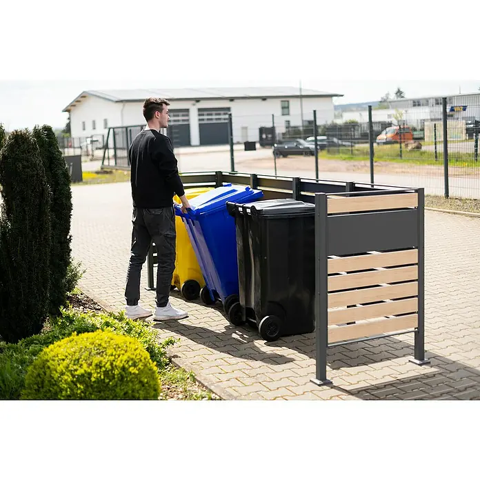 Westmann Mülltonnenbox Verkleidung Planum - 90 x 330 x 120 cm, Anthrazit/Holzoptik - Ein Mann schiebt eine blaue Mülltonne in eine Mülltonnenbox. Die Box ist aus grauem Metall und hellem Holz gefertigt.