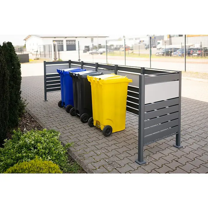 Westmann Mülltonnenbox Verkleidung Planum - 90 x 410 x 120 cm, Anthrazit/Grau - Drei Mülltonnen in Blau, Schwarz und Gelb stehen hinter einer grauen Sichtschutzwand auf Pflastersteinen.