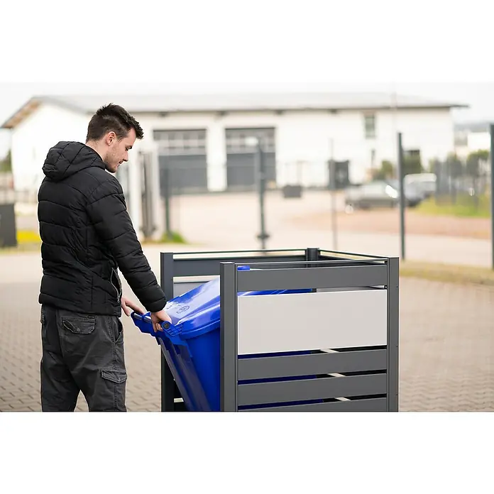 Westmann Mülltonnenbox Verkleidung Planum - 90 x 90 x 120 cm, Anthrazit/Grau - Ein Mann schiebt eine blaue Mülltonne in einen grauen Mülltonnenbox. Die Mülltonnenbox hat ein modernes Design mit weißen Akzenten.