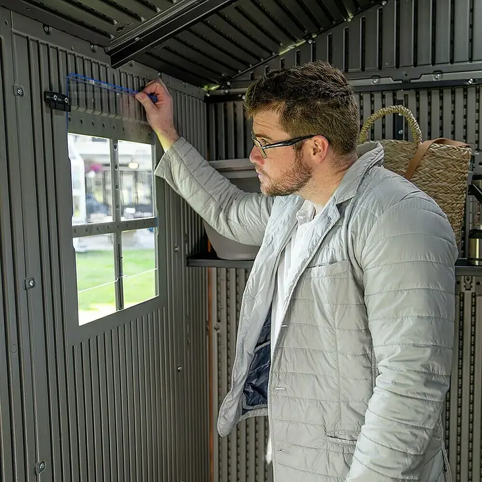Lifetime Gerätehaus Pluto - Außenbreite inkl. Dachüberstand: 356 cm, Außentiefe inkl. Dachüberstand: 213,1 cm, Kunststoff, Grau - Mann installiert eine klare Kunststoff-Fensterplatte in einem grauen Kunststoff-Gartenschuppen.
