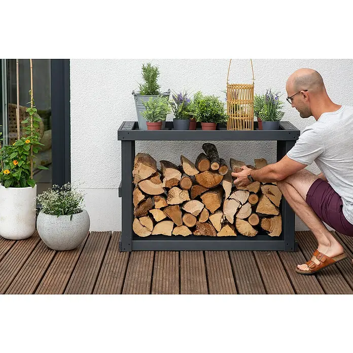 Westmann Kaminholzregal Stephan - 108 x 52 x 74 cm, Holz, Anthrazit - Ein graues Holzregal gefüllt mit Holzscheiten steht auf einer Holzterrasse, während ein Mann Holz einräumt. Auf dem Regal stehen dekorative Pflanzen.