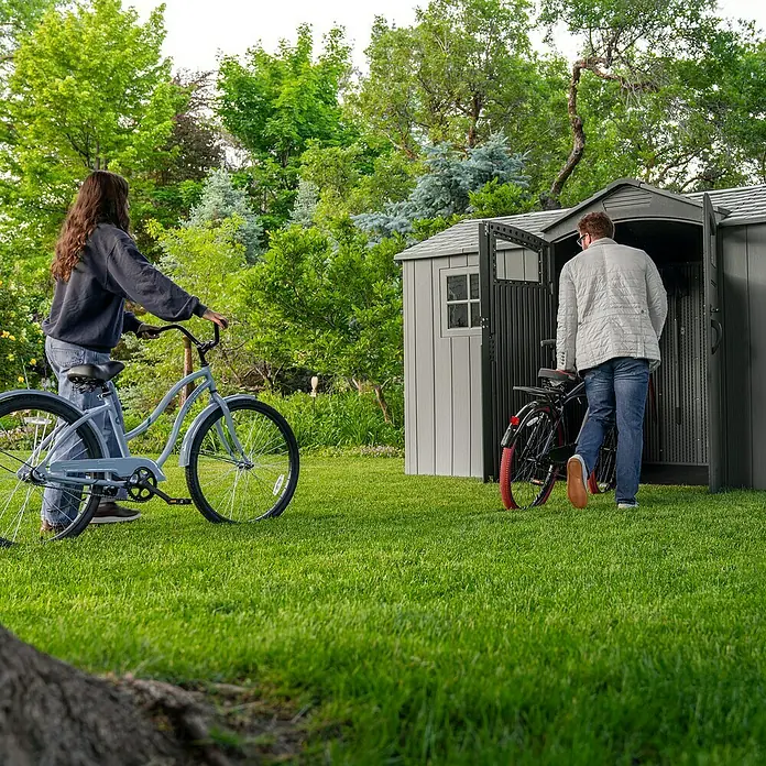 Lifetime Gerätehaus Pluto - Außenbreite inkl. Dachüberstand: 356 cm, Außentiefe inkl. Dachüberstand: 213,1 cm, Kunststoff, Grau - Grauer Gartenschuppen im Garten, in dem zwei Personen Fahrräder verstauen.