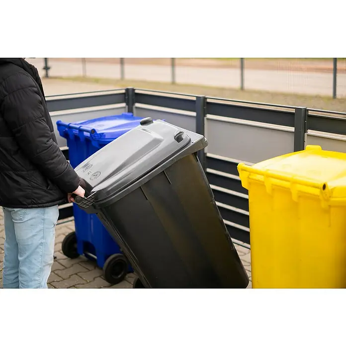 Westmann Mülltonnenbox Verkleidung Planum - 90 x 250 x 120 cm, Anthrazit/Grau - Eine Person zieht einen schwarzen Müllcontainer mit Rädern vor einem blauen und einem gelben Container.
