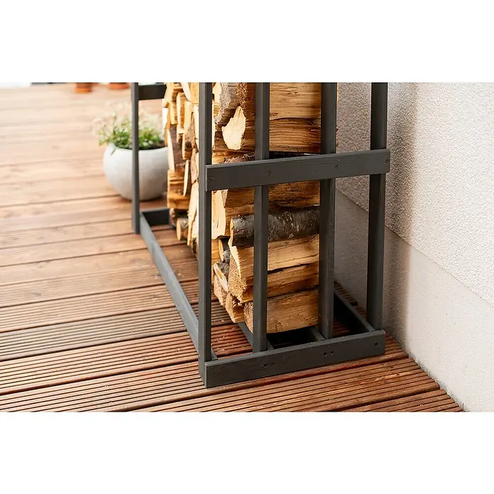 Westmann Kaminholzregal Harald - 108 x 52 x 106 cm, Holz, Anthrazit - Nahaufnahme eines dunklen Holzregals gefüllt mit Brennholz, das auf einer Holzterrasse steht, neben einer weißen Wand.