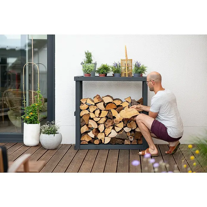 Westmann Kaminholzregal Harald - 108 x 52 x 106 cm, Holz, Anthrazit - Eine moderne Brennholzlagerung mit grauem Rahmen, gefüllt mit Holzscheiten und dekoriert mit Pflanzen. Ein Mann hockt daneben und hält Holz.