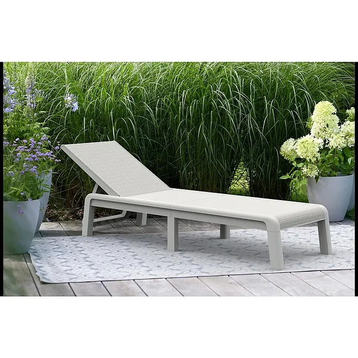 Keter Tumbona de jardín Mallorca - 58 x 187 x 29 cm, Blanco, Plástico, Respaldo regulable - Una elegante tumbona de exterior gris con respaldo ajustable se encuentra sobre una alfombra estampada, rodeada de exuberante vegetación y flores.