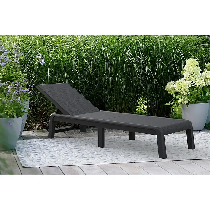 Keter Tumbona de jardín Mallorca - 58 x 187 x 29 cm, Grafito, Plástico, Respaldo regulable - Una tumbona de patio gris con respaldo ajustable se encuentra en un patio, con plantas verdes al fondo.