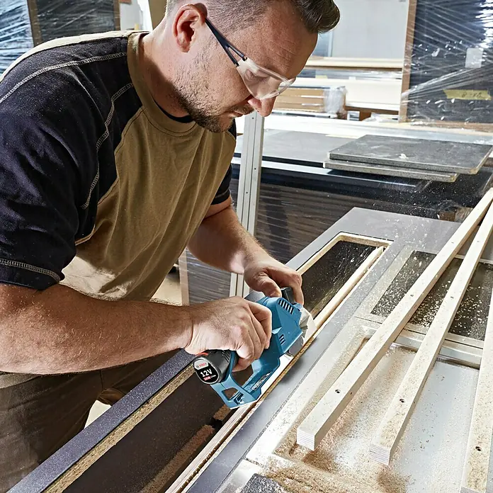 Bosch Professional Akku-Handhobel GHO 12V-20 - 12 V, Ohne Akku, 14.500 U/min - Ein Mann, der mit einer blauen Bosch 12V-Elektrohobelmaschine arbeitet, während er eine Schutzbrille trägt, um Holz zu bearbeiten.