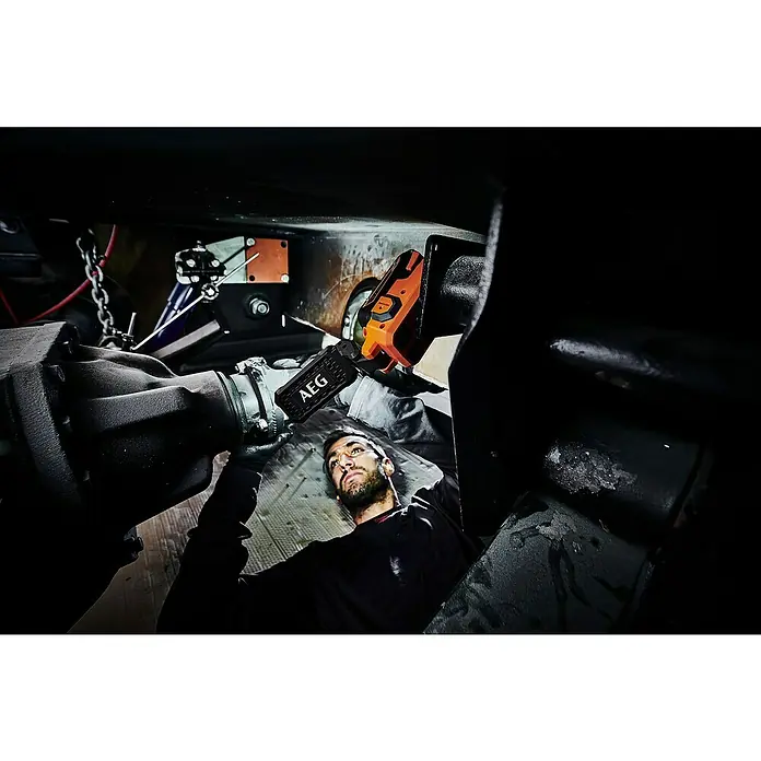AEG Pro 18V LED-Arbeitsleuchte BML 18 - 18 V, Ohne Akku, 850 lm, Aufhängehaken - Mann nutzt orange-schwarzen AEG Schlagschrauber zur Arbeit an dunklem Bauteil unter Maschine.