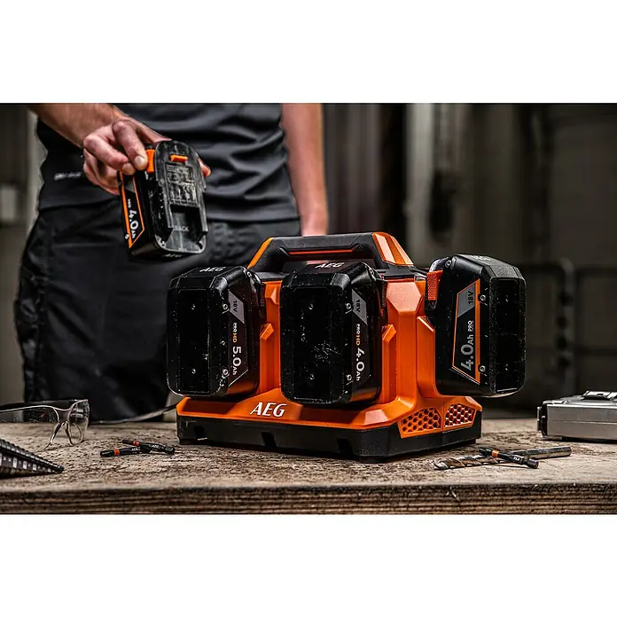 AEG Pro 18V Batterie-Ladegerät BL18C6 - 18 V, Ohne Akku, Ladestrom: 4 A - Orange-schwarzes AEG 18V Mehrfach-Akkuladegerät mit Akkus, einer wird von einer Person auf einer Werkbank eingesetzt.