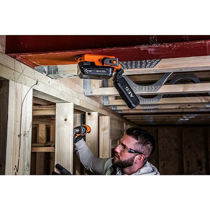 AEG Pro 18V LED-Arbeitsleuchte BML 18 - 18 V, Ohne Akku, 850 lm, Aufhängehaken - AEG 18V PRO HD 2.0Ah Akku mit LED-Arbeitsleuchte, orange und schwarz, beleuchtet Baustellenbereich. Ein Mann bohrt im Hintergrund.