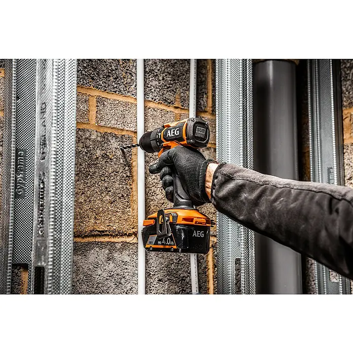 AEG Pro 18V Akku-Bohrschrauber BSB 18SBL2-0 - 18 V, Ohne Akku, 65 Nm - Orange-schwarze AEG Akkubohrmaschine, von einer behandschuhten Hand gehalten, bohrt in eine graue Ziegelwand neben Metallprofilen.
