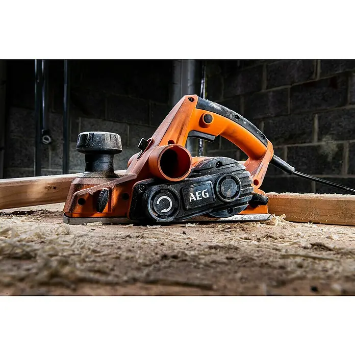 AEG Elektrohobel PL700 700 W - 700 W, Hobeltiefe: 0 mm - 2 mm, Leerlaufdrehzahl: 17.000 U/min - Ein orange-schwarzer AEG Elektrohobel, bedeckt mit Sägespänen, ruht auf Holzspänen auf einer Holzoberfläche neben einem Brett in einer Werkstatt.