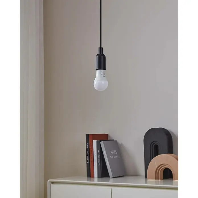 Eglo connect.z Smart-LED-Lampe Zigbee RGB/CCT - E27, Dimmbarkeit: Dimmbar, RGBW, 806 lm, 9 W - Eine einzelne Glühbirne hängt an einem schwarzen Kabel vor einer Wand, beleuchtet Bücher und Deko auf einem weißen Tisch.