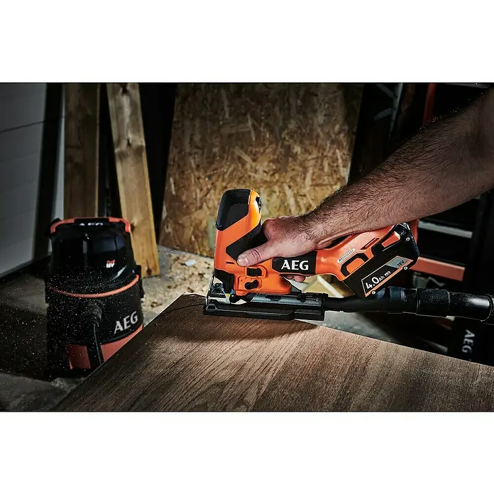 AEG Pro 18V Akku-Stichsäge BST 18BSBL - 18 V, Ohne Akku, Leerlaufhubzahl: 900 /min - 3.200 /min - Orange-schwarze AEG Stichsäge schneidet Holz, daneben ein Staubsauger in einer Werkstatt.