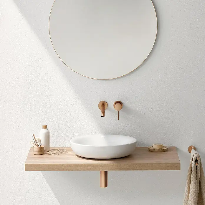 Grohe Essence Waschtischarmatur M - Wandmontage, Warm Sunset, Matt - Ein modernes Badezimmer-Waschbecken mit runder Spiegel, hellen Holztönen und stilvollen Armaturen, die eine elegante und minimalistische Atmosphäre schaffen.