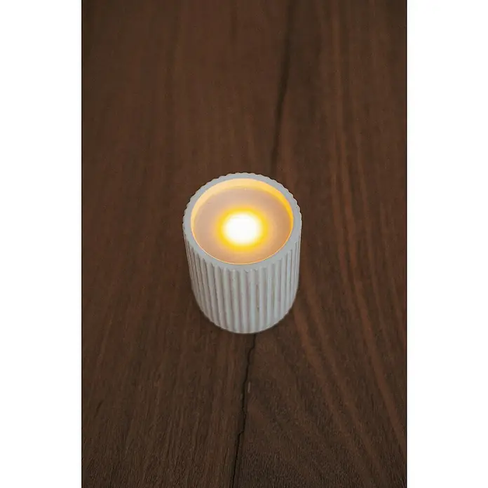 New Garden Vela LED para exteriores Dolo - 10 cm, Blanco - Primer plano de una vela de té LED acanalada sobre una mesa de madera oscura, que emite un brillo cálido y suave.