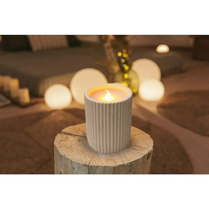 New Garden Vela LED para exteriores Dolo - 10 cm, Blanco - Vela LED decorativa en un recipiente de cerámica acanalada, colocada sobre un taburete de madera, rodeada de una iluminación ambiental suave.