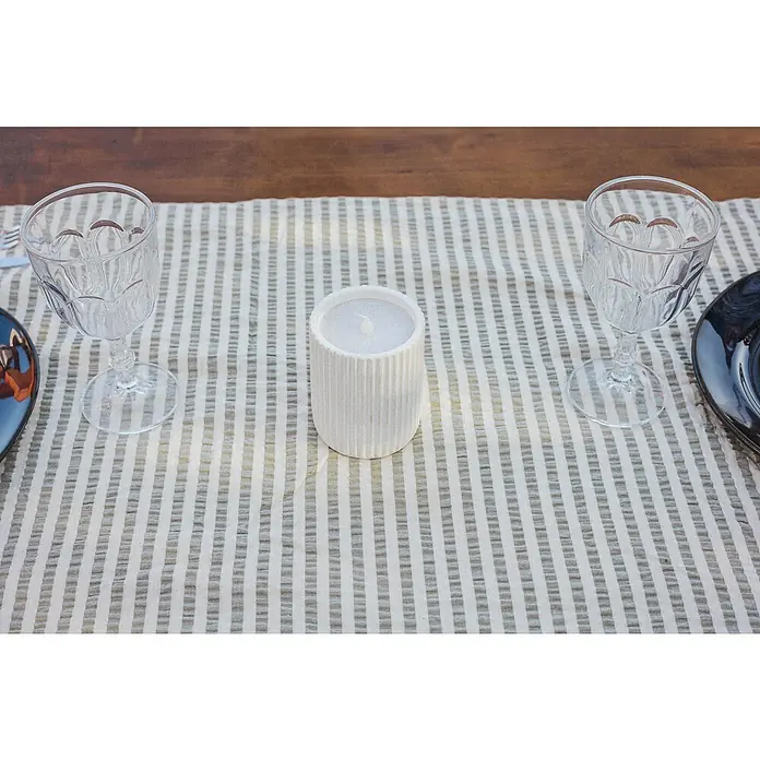 New Garden Vela LED para exteriores Dolo - 10 cm, Blanco - Una vela blanca sobre una mesa con dos copas de vino decorativas y un camino de mesa a rayas.