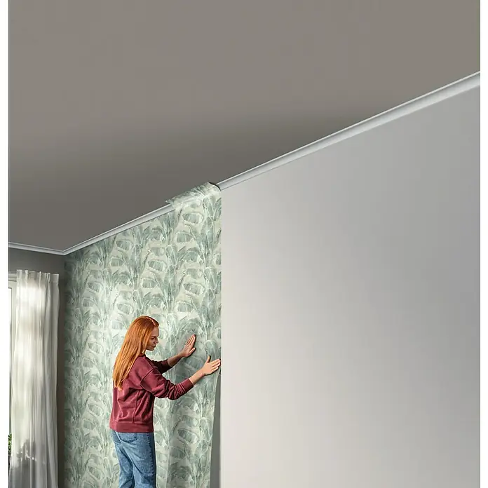 Tesa Rasch Strukturtapete Cuba Blatt - Selbstklebend, Grün, Floral, 6 x 0,53 m - Eine Frau bringt eine stilvolle Blattmuster-Tapete an einer Wand in einem Raum an, die dem Interieur einen Hauch von Natur verleiht.