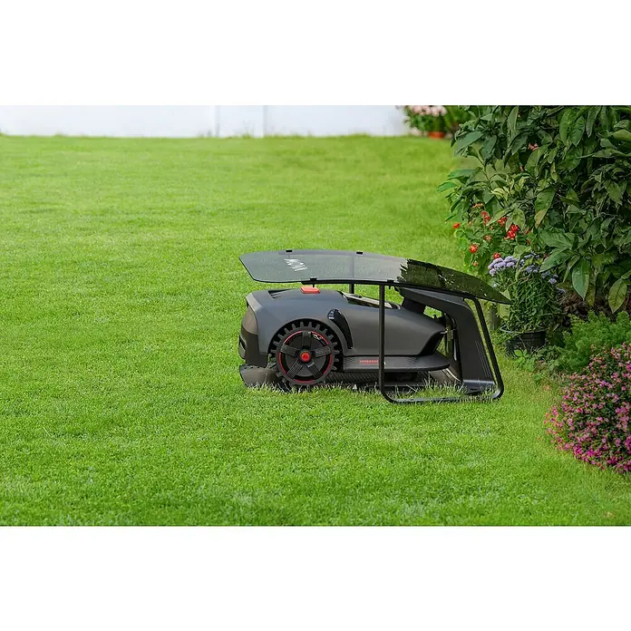 Mova Mähroboter-Garage - L x B x H: 785,5 x 590 x 378,4 mm, Mova Mähroboter 1000, MOVA 600 und 600 Kit - Ein Rasenmähroboter im Garten. Der Mähroboter ist grau mit roten Akzenten und steht auf einem grünen Rasen, neben einem Busch mit bunten Blumen.