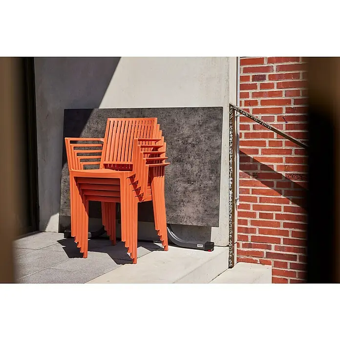 Siena Garden Urban Lights Gartenmöbel-Set Lane Sessel/Paco Tisch - 5 -tlg., Polypropylen, Brick/Anthrazit - Eine Nahaufnahme eines Stapels oranger Plastikstühle mit Armlehnen vor einer Betonwand und einer Backsteinmauer. Die Stühle sind für den Außenbereich geeignet.