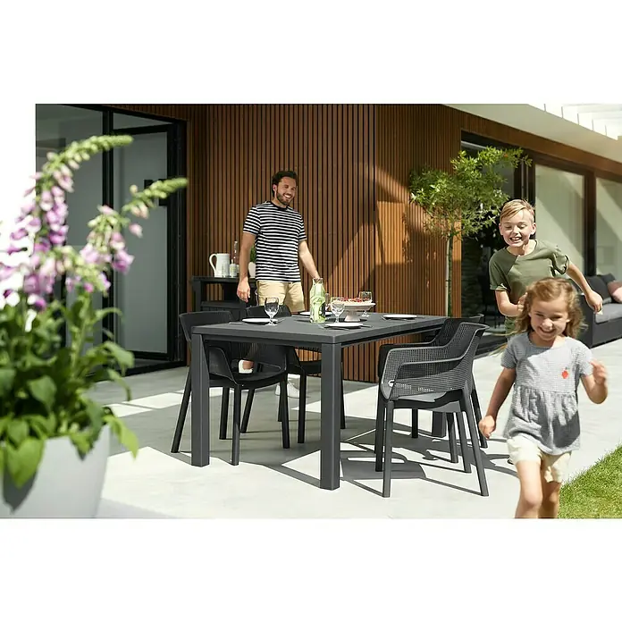 Keter Mesa de jardín Julie - L x An x Al: 149 x 92 x 75 cm, Polipropileno, Grafito - Imagen de una mesa de jardín gris con cuatro sillas en un patio con vajilla puesta. Niños jugando al fondo.