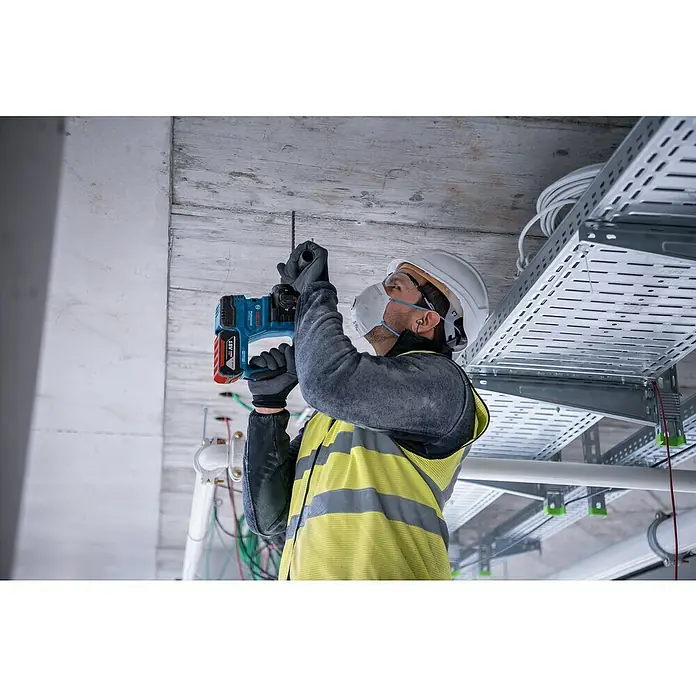 Bosch Professional AMPShare 18V Akku-Bohrhammer GBH 18V-22 - 18 V, Ohne Akku, 1,9 J - Ein Bauarbeiter mit Schutzhelm und Maske bohrt mit einer Bosch Akku-Bohrmaschine in eine Betondecke. Im Hintergrund sind Kabelkanäle zu sehen.