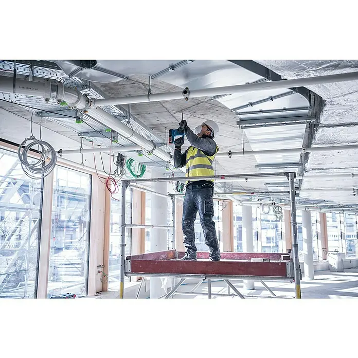 Bosch Professional AMPShare 18V Akku-Bohrhammer PRO GBH 18V-22 - 18 V, 2 Akkus, 4 Ah, 1,9 J - Ein Bauarbeiter auf einem Gerüst befestigt eine Leitung an der Decke mit einem blauen Bohrhammer. Er trägt Sicherheitsausrüstung wie Helm und Warnweste.