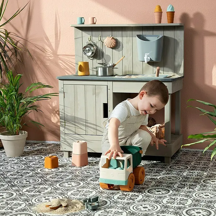 Muddy Buddy Matschküche Mud Expert80 x 40 x 90 cm, Holz, Warmgrau - Ein Junge spielt mit einem Spielzeug-LKW und Zubehör vor einer hölzernen Spielküche im Freien.