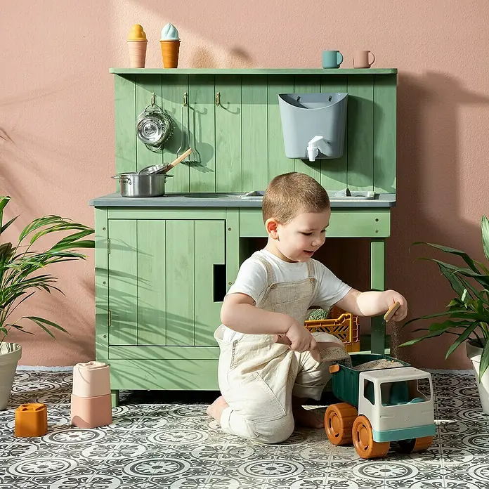 Muddy Buddy Matschküche Mud Expert80 x 40 x 90 cm, Holz, Eukalyptus - Ein Kind spielt mit einem Spielzeug-Lastwagen vor einer grünen Spielküche, die mit Töpfen und Spielzeug-Eis dekoriert ist.