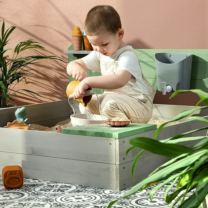 Muddy Buddy Sandkasten Cozy House - 100 x 100 x 60 cm, Holz, Warmgrau/ Eukalyptus - Ein Kind spielt in einem Sandkasten mit Spielzeug, darunter ein Dinosaurier und Eistüten. Der Sandkasten ist aus grauem Holz gefertigt.