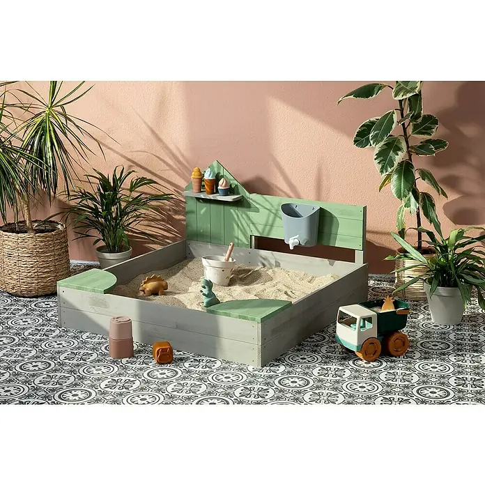 Muddy Buddy Sandkasten Cozy House - 100 x 100 x 60 cm, Holz, Warmgrau/ Eukalyptus - Ein detailreiches Bild eines Sandkastens mit Spielzeug, umgeben von Zimmerpflanzen auf einem gemusterten Boden.