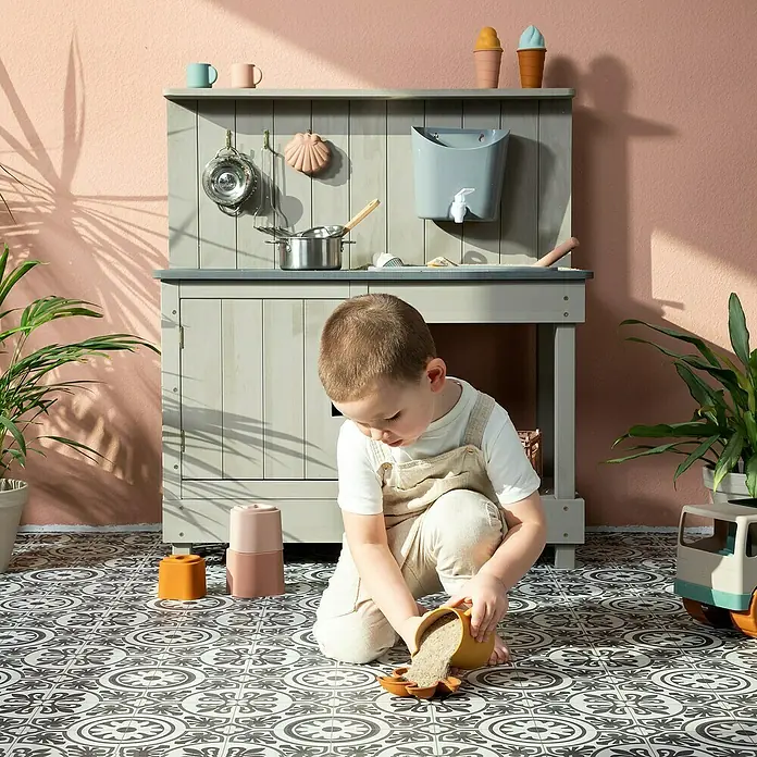 Muddy Buddy Matschküche Mud Expert - 80 x 40 x 90 cm, Holz, Warmgrau - Ein Kind spielt mit dem hölzernen Küchenspielzeug und Sandformen auf dem Boden. Die Spielküche aus Holz steht im Hintergrund.