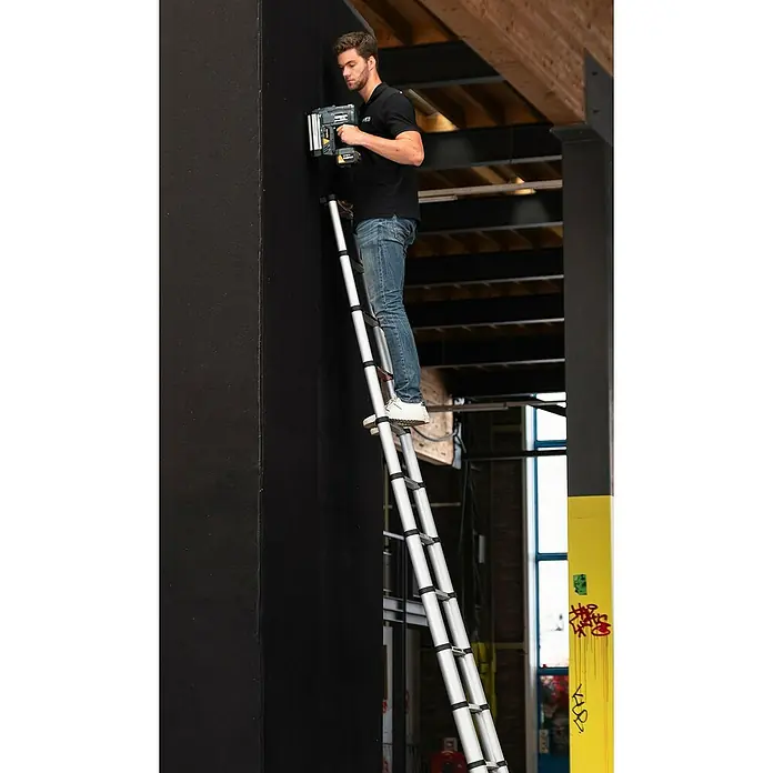 Batavia Teleskopleiter - Anzahl Tritte: 14 Stk., Arbeitshöhe: 515 cm, Belastbarkeit: 150 kg, Aluminium - Ein Mann, der auf einer Leiter steht und mit einem Nagler an einer schwarzen Wand arbeitet, präsentiert das Werkzeug in Aktion.