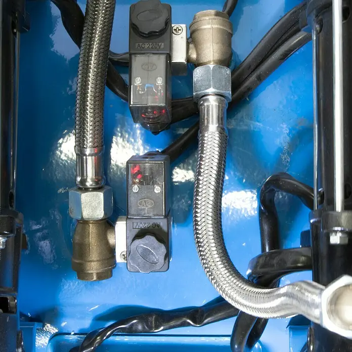 Güde Flüsterkompressor 375/8/100 ED-Silent - 2.200 W, Max. Druck: 8 bar, 100 l, Schmierung: Ohne - Zwei Industriesolenoidventile mit aktiven roten Anzeigen, verbunden mit geflochtenen Metallschläuchen und Messingarmaturen auf blauer Platte.