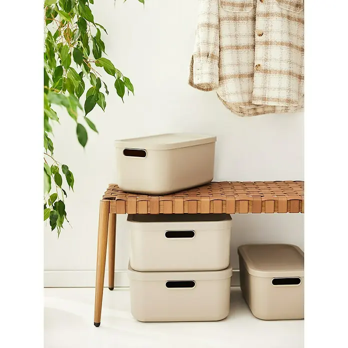 Rotho Aufbewahrungsbox Albula - L x B x H: 39,5 x 23,5 x 17,7 cm, Kunststoff, Cappuccino, 12 l - Beige Aufbewahrungsboxen mit Griffen, gestapelt auf und neben einer Bank aus geflochtenem Leder.