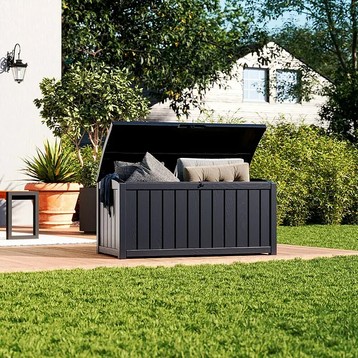 Keter Gartenbox Glenwood - L x B x H: 128 x 65 x 61 cm, Kunststoff, Anthrazit - Eine offene, schwarze Gartenbox gefüllt mit Kissen auf einer Terrasse, umgeben von einem gepflegten Garten.
