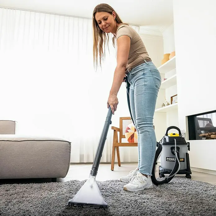 PowerPlus Aspirador en seco y húmedo eléctrico POWX326 - 1.600 W, Volumen de recipiente: 20 l, Con bolsa - Una mujer está usando un limpiador de alfombras POWERPLUS con un accesorio de manguera para limpiar una alfombra gris en su sala de estar.