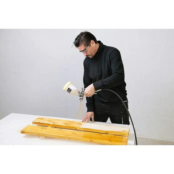 PowerPlus Pistola de pintura neumática POWAIR0109 - Presión de funcionamiento: 3 bar, Volumen de recipiente: 600 ml - Un hombre con gafas de seguridad rocía pintura sobre tablas de madera con un rociador de pintura para el bricolaje.