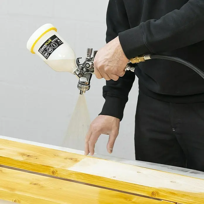 PowerPlus Pistola de pintura neumática POWAIR0109 - Presión de funcionamiento: 3 bar, Volumen de recipiente: 600 ml - Una persona rocía pintura sobre una tabla de madera con una pistola de pintura para recubrirla. La pistola está equipada con un recipiente de pintura.
