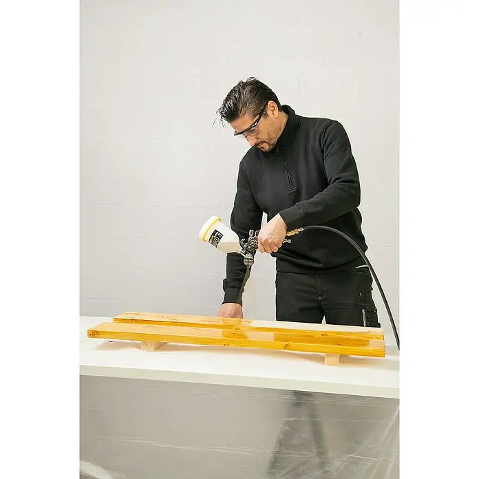 PowerPlus Pistola de pintura neumática POWAIR0109 - Presión de funcionamiento: 3 bar, Volumen de recipiente: 600 ml - Hombre barnizando madera con pistola de pulverización de pintura. Gafas de seguridad y ropa de trabajo.