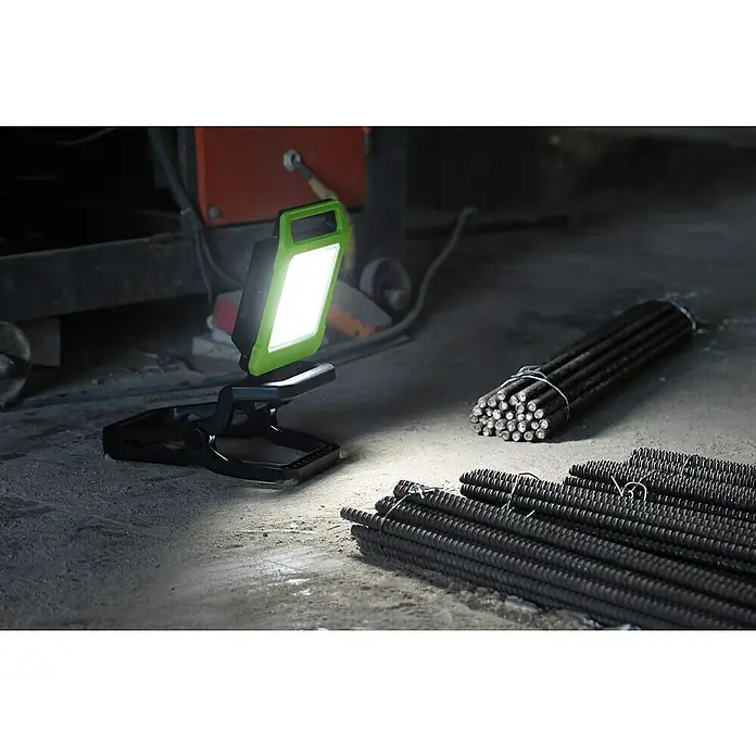 Profi Depot LED-Akkustrahler 2 in 1 - 20 W, 5.600 lm, 6.500 K, 12,7 x 12,7 x 330 cm, IP54 - Akkubetriebene Arbeitsleuchte mit grünem Rahmen, die eine Baustelle mit Bewehrungsstäben beleuchtet.