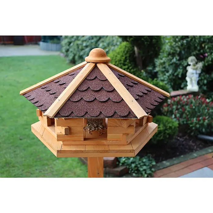 Promadino Vogelhaus Knusperhäuschen - Holz, Natur/Braun, Ausstattung: Ohne Vogelhausständer, Mit Silo - Nahaufnahme eines hölzernen Vogelhauses mit braunem Dach und Futter darin, aufgestellt in einem Garten mit grüner Umgebung.