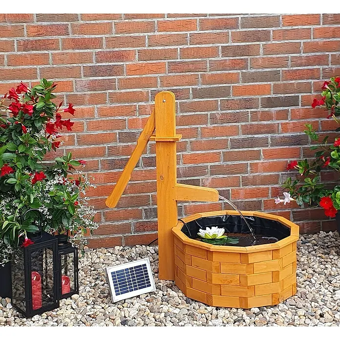 Promadino Standbrunnen - Ø x H: 64 x 101 cm, Solarpumpe - Gartenszene mit einem dekorativen Holzbrunnen, gespeist durch eine Solarzelle, umgeben von Steinen und roten Blüten.
