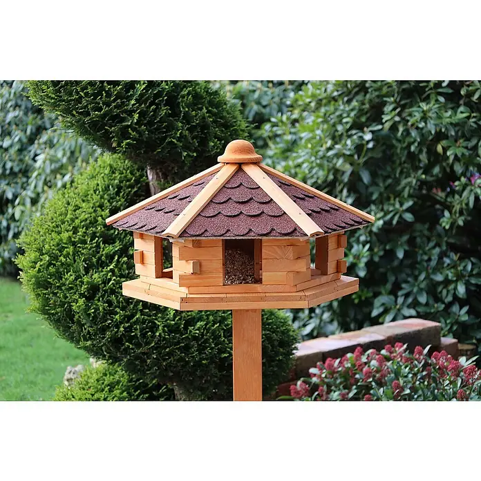 Promadino Vogelhaus Knusperhäuschen - Holz, Natur/Braun, Ausstattung: Ohne Vogelhausständer, Mit Silo - Ein detailreiches Vogelhaus aus Holz mit einem braunen Schindeldach, das im Garten zwischen üppigem Grün steht. Es ist mit Vogelfutter gefüllt.