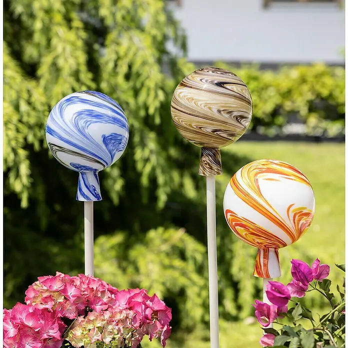 Windhager Glaskugel - Durchmesser: 15 cm, Braun - Nahaufnahme von drei dekorativen Gartenkugeln in Blau, Braun und Orange mit Blumen im Vordergrund, die einen Hauch von Eleganz verleihen.
