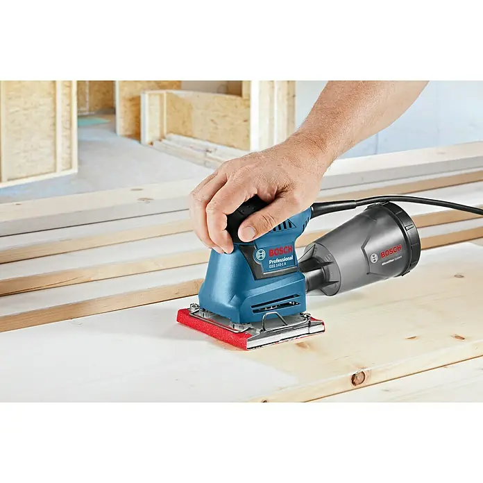 Bosch Professional Lijadora orbital eléctrica GSS 140-1A - 180 W, Número de oscilaciones marcha en vacío: 24.000 r.p.m. - Se está utilizando una lijadora orbital Bosch Professional GSS 140-1 A sobre una superficie de madera. La herramienta es azul y tiene un depósito de polvo gris.