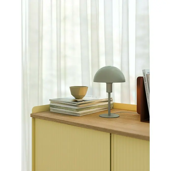 Nordlux Lámpara de sobremesa Ellen mini - 40 W, L x An x Al: 16 x 16 x 25 cm, Verde, E14 - Lámpara seta verde salvia sobre mueble amarillo acanalado, con revistas y cuenco.