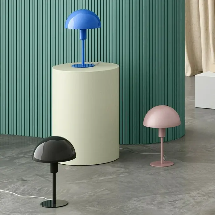 Nordlux Lámpara de sobremesa Ellen mini - 40 W, L x An x Al: 16 x 16 x 25 cm, Verde, E14 - Tres lámparas de hongo en azul, gris y rosa están sobre una superficie cilíndrica beige y en un suelo gris sobre un fondo verde.